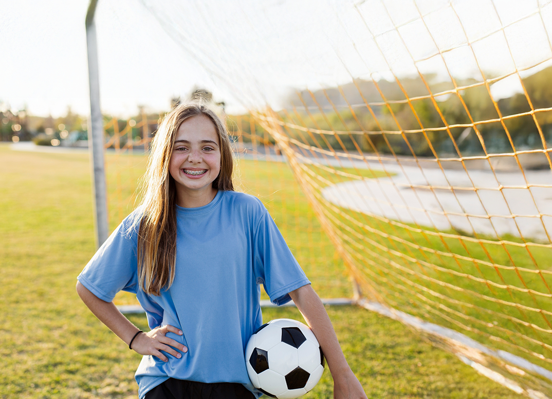 Girl with traditional braces smiling while holding a soccer ball next to the goal